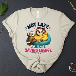 May include: Beige t-shirt with a cartoon sloth wearing sunglasses and a yellow hoodie, relaxing on a blue beanbag chair. The text reads "NOT LAZY, JUST SAVING ENERGY." The sloth holds a coffee mug.