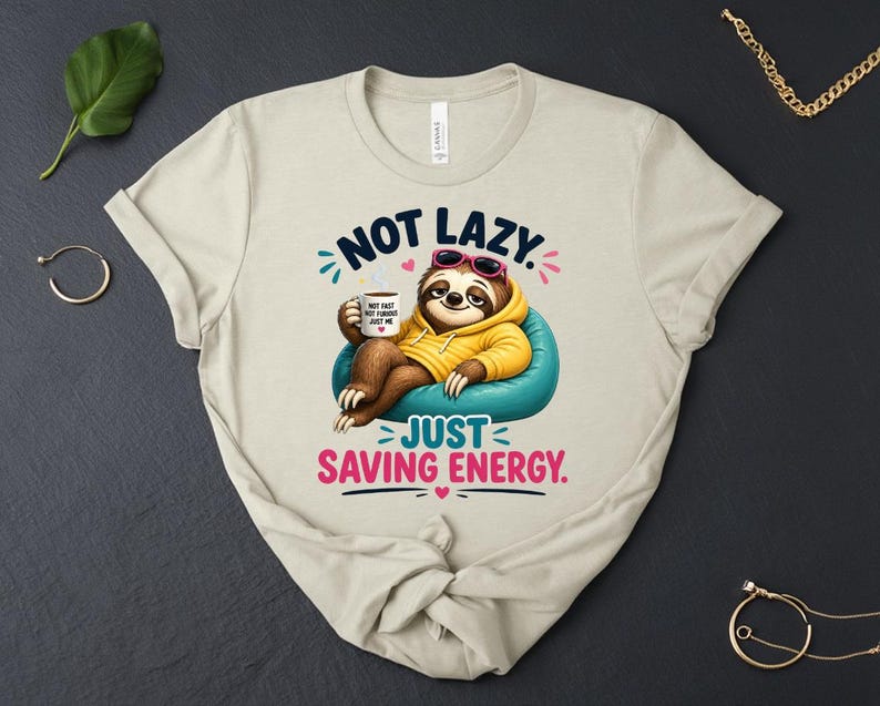 May include: Beige t-shirt with a cartoon sloth wearing sunglasses and a yellow hoodie, relaxing on a blue beanbag chair. The text reads "NOT LAZY, JUST SAVING ENERGY." The sloth holds a coffee mug.