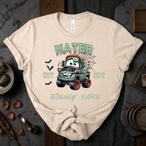 Może przedstawiać: Beżowa koszulka z grafiką Mater z filmu Auta Disneya w stylu zombie. Projekt zawiera napisy "MATER", "EST 1977" i "Disney Cars" w kolorze zielonym, a także nietoperze i pajęczynę.