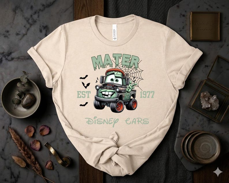 Może przedstawiać: Beżowa koszulka z grafiką Mater z filmu Auta Disneya w stylu zombie. Projekt zawiera napisy "MATER", "EST 1977" i "Disney Cars" w kolorze zielonym, a także nietoperze i pajęczynę.