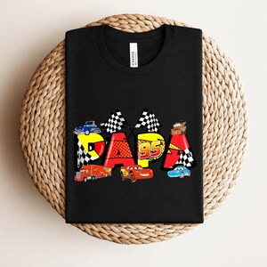 Puede incluir: Camiseta negra con la palabra "DAPA" en un dise&ntilde;o colorido con coches de carreras y el n&uacute;mero 95. El dise&ntilde;o incluye banderas a cuadros y varias ilustraciones de coches, creando un aspecto l&uacute;dico y vibrante.