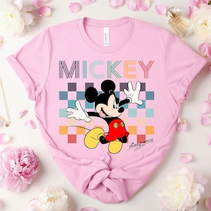 May include: A light pink t-shirt featuring a colorful graphic of Mickey Mouse. The word "MICKEY" is printed across the top in a striped font. Mickey Mouse is depicted with his signature black ears, red shorts, yellow shoes, and white gloves.