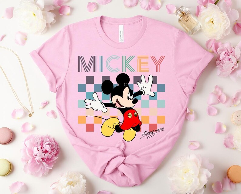 May include: A light pink t-shirt featuring a colorful graphic of Mickey Mouse. The word "MICKEY" is printed across the top in a striped font. Mickey Mouse is depicted with his signature black ears, red shorts, yellow shoes, and white gloves.