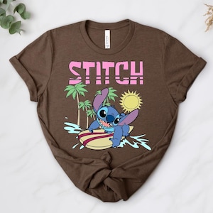 May include: Brown t-shirt featuring a graphic of Stitch surfing on a surfboard. The word "STITCH" is printed in pink above the character. Palm trees and a yellow sun are also included in the design.