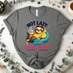 May include: Grey t-shirt featuring a cartoon sloth in sunglasses and a yellow hoodie, holding a coffee cup. The text says "NOT LAZY. JUST SAVING ENERGY."