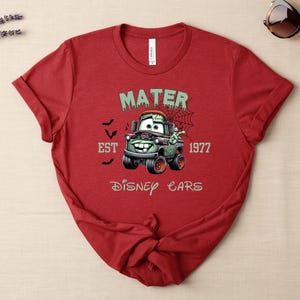 Może przedstawiać: Czerwona koszulka z motywem zombie Mater z filmu Auta Disneya. Projekt zawiera tekst "MATER", "EST 1977" i "Disney Cars". Koszulka ma kr&oacute;tki rękaw i okrągły dekolt.