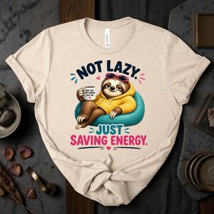 May include: Beige t-shirt with a cartoon sloth wearing sunglasses and a yellow hoodie, relaxing on a blue beanbag chair. The text reads "NOT LAZY. JUST SAVING ENERGY." The sloth holds a mug that says "NOT FAST, NOT FURIOUS, JUST ME."