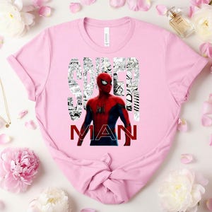 Peut inclure: Un t-shirt rose clair &agrave; manches courtes avec un graphisme du personnage Spider-Man en costume rouge et bleu. Le mot SPIDER est rempli de panneaux de bande dessin&eacute;e en noir et blanc, et le mot MAN est imprim&eacute; en lettres rouges grasses.