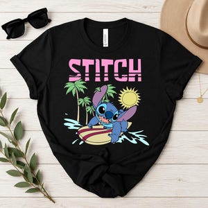 May include: Black t-shirt featuring a graphic of Stitch surfing on a surfboard, with palm trees and a sun in the background. The word "STITCH" is printed in pink block letters above the graphic.