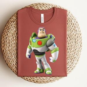 May include: A rust-colored t-shirt featuring a graphic of Buzz Lightyear from Toy Story. The character is depicted in his signature green, white, and purple spacesuit. The shirt is folded on a woven, circular surface.