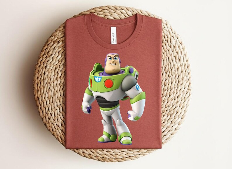 May include: A rust-colored t-shirt featuring a graphic of Buzz Lightyear from Toy Story. The character is depicted in his signature green, white, and purple spacesuit. The shirt is folded on a woven, circular surface.