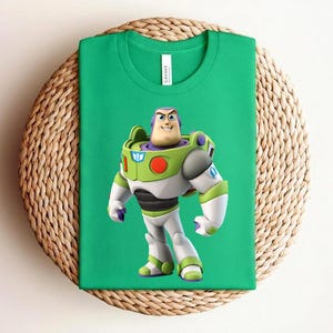 May include: A green t-shirt featuring a colorful graphic of Buzz Lightyear from Toy Story. The character is depicted in his signature space ranger uniform, with white, green, and purple accents. The shirt is displayed on a woven, circular surface.