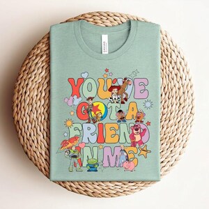 May include: A light green t-shirt with the phrase "You've Got a Friend in Me" in colourful letters, with cartoon characters. The shirt has a crew neck and short sleeves. The design includes characters from the Toy Story film.