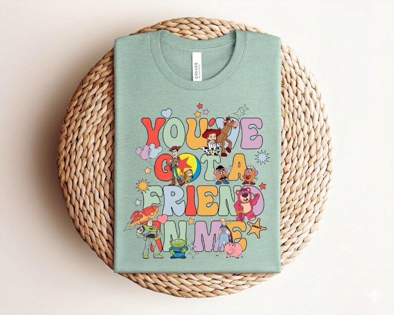 May include: A light green t-shirt with the phrase "You've Got a Friend in Me" in colourful letters, with cartoon characters. The shirt has a crew neck and short sleeves. The design includes characters from the Toy Story film.
