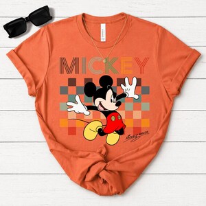 May include: Orange t-shirt featuring Mickey Mouse with the word "MICKEY" in a retro font. Mickey is in a dynamic pose against a colorful checkered background. The shirt includes the signature "Mickey Mouse" in script.