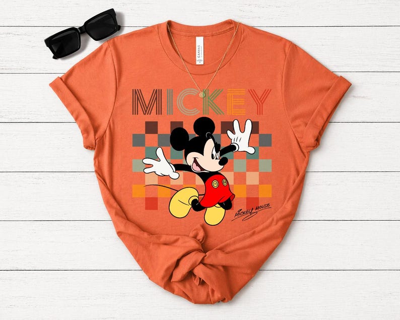 May include: Orange t-shirt featuring Mickey Mouse with the word "MICKEY" in a retro font. Mickey is in a dynamic pose against a colorful checkered background. The shirt includes the signature "Mickey Mouse" in script.