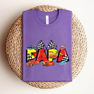 Puede incluir: Una camiseta morada con la palabra "PAPA" en un dise&ntilde;o colorido y caricaturesco con coches de carreras y banderas a cuadros. El n&uacute;mero "95" tambi&eacute;n est&aacute; incluido en el dise&ntilde;o.