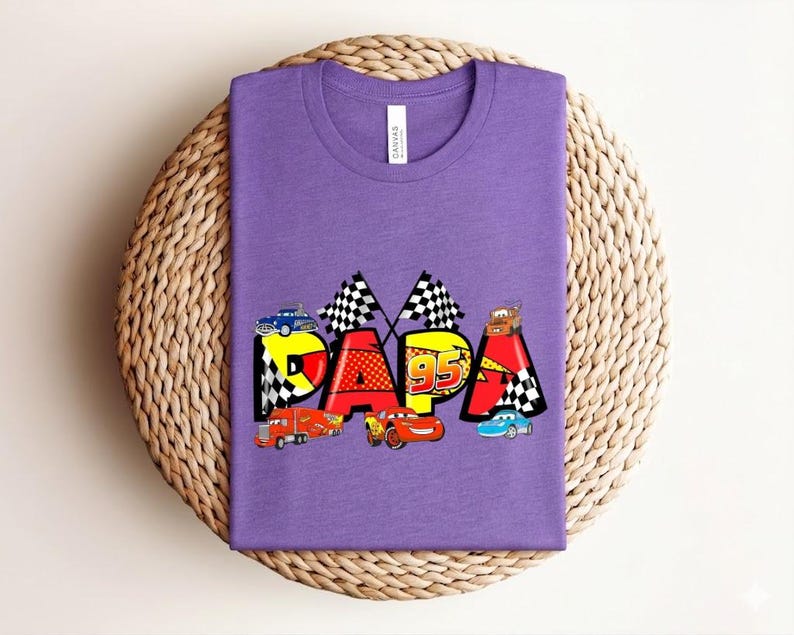 Puede incluir: Una camiseta morada con la palabra "PAPA" en un dise&ntilde;o colorido y caricaturesco con coches de carreras y banderas a cuadros. El n&uacute;mero "95" tambi&eacute;n est&aacute; incluido en el dise&ntilde;o.