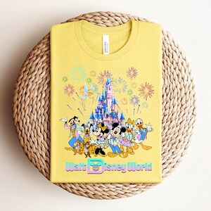 Może przedstawiać: Ż&oacute;łta koszulka z kolorową grafiką postaci Disneya, zamkiem i fajerwerkami. Tekst "Walt Disney World" jest wyświetlany w gradiencie. Koszulka jest złożona na tkanej, okrągłej powierzchni.