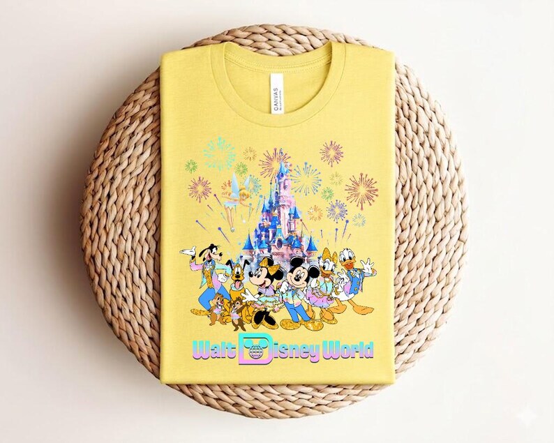 Może przedstawiać: Ż&oacute;łta koszulka z kolorową grafiką postaci Disneya, zamkiem i fajerwerkami. Tekst "Walt Disney World" jest wyświetlany w gradiencie. Koszulka jest złożona na tkanej, okrągłej powierzchni.