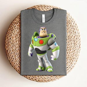 May include: A gray t-shirt featuring a colorful graphic of Buzz Lightyear from Toy Story. The character is depicted in his signature green, white, and purple spacesuit. The shirt is folded on a woven, circular mat.