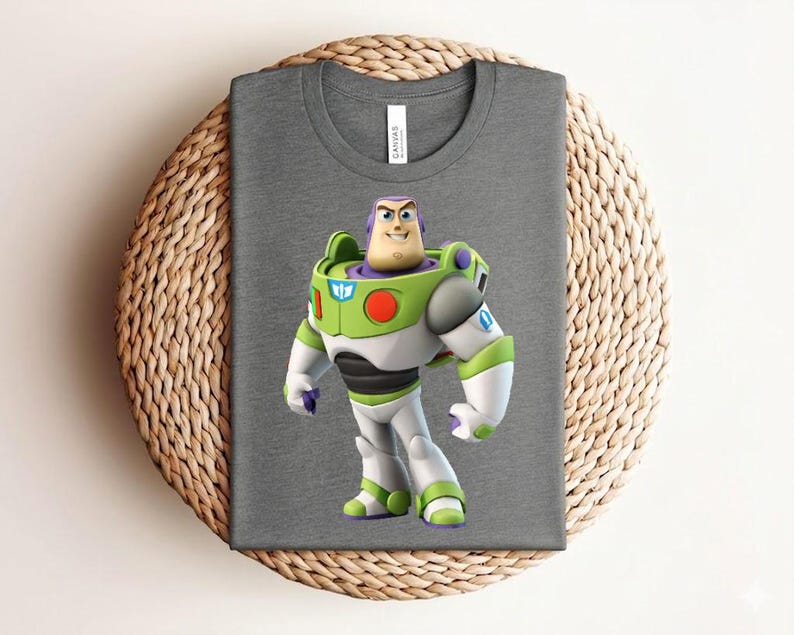 May include: A gray t-shirt featuring a colorful graphic of Buzz Lightyear from Toy Story. The character is depicted in his signature green, white, and purple spacesuit. The shirt is folded on a woven, circular mat.