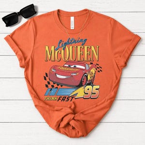 May include: Orange t-shirt with a Lightning McQueen design from the Cars film. The graphic includes the text "Lightning McQueen", the number "95", and the phrase "Think Fast". Black sunglasses and a gold necklace are also visible.