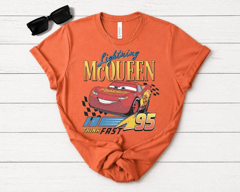 May include: Orange t-shirt with a Lightning McQueen design from the Cars film. The graphic includes the text "Lightning McQueen", the number "95", and the phrase "Think Fast". Black sunglasses and a gold necklace are also visible.