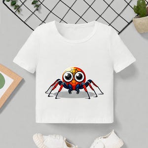 Pu&ograve; includere: T-shirt bianca a maniche corte con una grafica di ragno dei cartoni animati. Il ragno ha un corpo rosso, arancione e giallo con grandi occhi e zampe nere e rosse. Un paio di scarpe da ginnastica bianche sono nella parte inferiore dell'immagine.