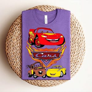 May include: A purple t-shirt featuring a graphic of the Cars movie characters. The design includes Lightning McQueen, Mater, and a yellow car, with the word "Cars" in a stylized font. The shirt is displayed on a woven surface.