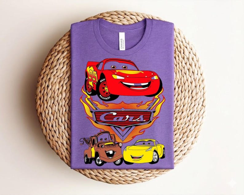 May include: A purple t-shirt featuring a graphic of the Cars movie characters. The design includes Lightning McQueen, Mater, and a yellow car, with the word "Cars" in a stylized font. The shirt is displayed on a woven surface.