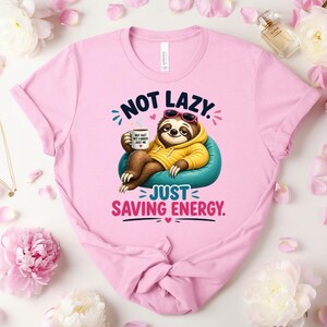 May include: Pale pink t-shirt with a cartoon sloth wearing sunglasses and a yellow hoodie, relaxing on a blue beanbag chair. The text reads "NOT LAZY. JUST SAVING ENERGY."