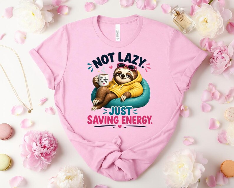 May include: Pale pink t-shirt with a cartoon sloth wearing sunglasses and a yellow hoodie, relaxing on a blue beanbag chair. The text reads "NOT LAZY. JUST SAVING ENERGY."