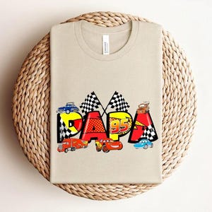 Puede incluir: Una camiseta beige con la palabra "DAPA" en letras coloridas de estilo caricatura. Las letras est&aacute;n decoradas con coches de carreras y banderas a cuadros. El n&uacute;mero "95" tambi&eacute;n est&aacute; presente en el dise&ntilde;o.