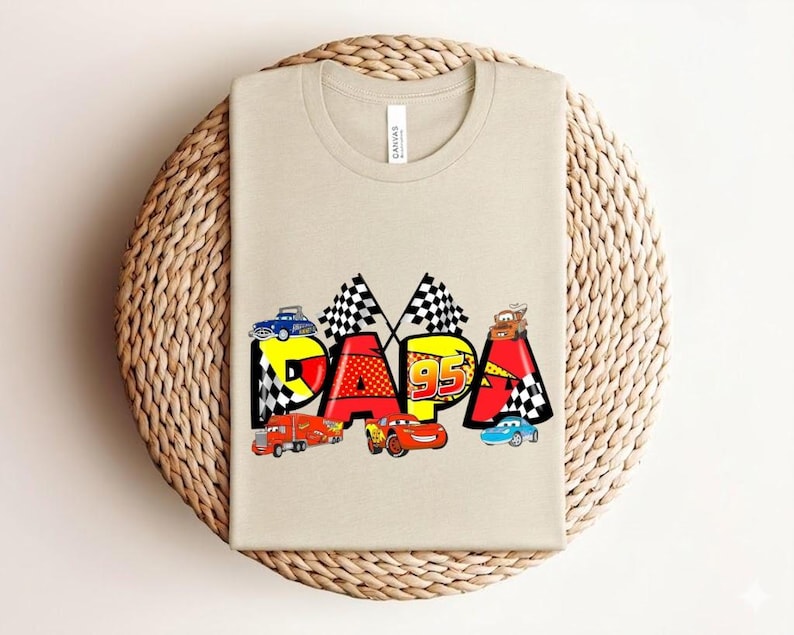 Puede incluir: Una camiseta beige con la palabra "DAPA" en letras coloridas de estilo caricatura. Las letras est&aacute;n decoradas con coches de carreras y banderas a cuadros. El n&uacute;mero "95" tambi&eacute;n est&aacute; presente en el dise&ntilde;o.