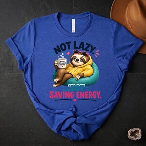 May include: Royal blue t-shirt featuring a cartoon sloth in sunglasses and a yellow hoodie, holding a coffee cup. The text on the shirt says "Not Lazy, Just Saving Energy." The sloth is on a teal beanbag chair.