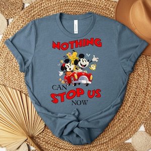 Puede incluir: Una camiseta azul con una imagen de dibujos animados de Mickey y Minnie Mouse en un coche rojo, con Pluto y un p&aacute;jaro amarillo. El texto "NOTHING CAN STOP US NOW" es rojo. La camiseta est&aacute; sobre una estera tejida con un sombrero marr&oacute;n.