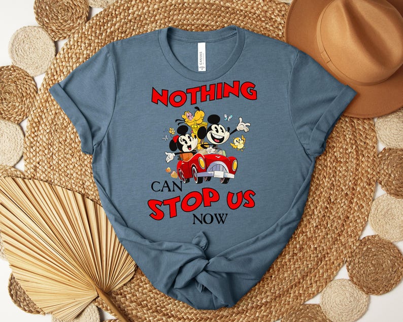 Puede incluir: Una camiseta azul con una imagen de dibujos animados de Mickey y Minnie Mouse en un coche rojo, con Pluto y un p&aacute;jaro amarillo. El texto "NOTHING CAN STOP US NOW" es rojo. La camiseta est&aacute; sobre una estera tejida con un sombrero marr&oacute;n.