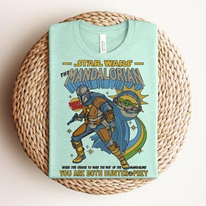 K&ouml;nnte beinhalten: Ein hellgr&uuml;nes T-Shirt mit einer Grafik von "The Mandalorian". Das Design enth&auml;lt den Text "Star Wars The Mandalorian" und eine Illustration des Mandalorianers und Grogu. Das Shirt hat auch den Text "When you choose to walk the way of the Mandalorian, you are both hunter & prey."