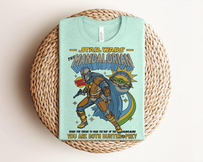 K&ouml;nnte beinhalten: Ein hellgr&uuml;nes T-Shirt mit einer Grafik von "The Mandalorian". Das Design enth&auml;lt den Text "Star Wars The Mandalorian" und eine Illustration des Mandalorianers und Grogu. Das Shirt hat auch den Text "When you choose to walk the way of the Mandalorian, you are both hunter & prey."