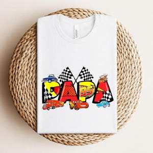 Puede incluir: Camiseta blanca con la palabra "PAPA" en un dise&ntilde;o colorido con coches de carreras y banderas a cuadros. El n&uacute;mero "95" tambi&eacute;n est&aacute; incluido en el dise&ntilde;o. La camiseta est&aacute; doblada sobre una superficie tejida.