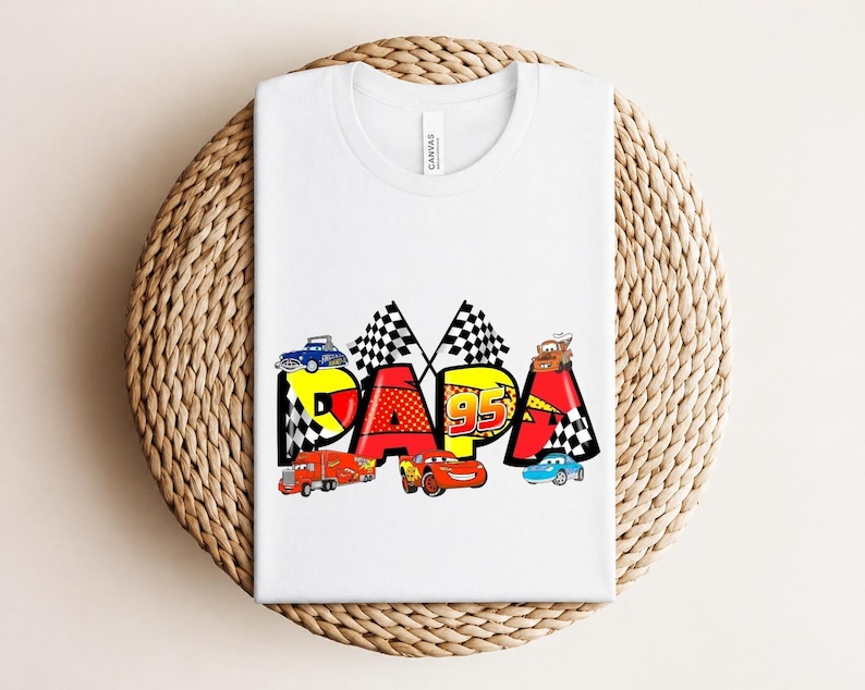 Puede incluir: Camiseta blanca con la palabra "PAPA" en un dise&ntilde;o colorido con coches de carreras y banderas a cuadros. El n&uacute;mero "95" tambi&eacute;n est&aacute; incluido en el dise&ntilde;o. La camiseta est&aacute; doblada sobre una superficie tejida.