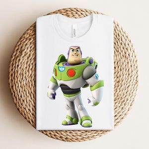 May include: White t-shirt featuring a colorful graphic of Buzz Lightyear from Toy Story. The character is depicted in his signature green, white, and purple spacesuit. The shirt is folded on a woven, circular surface.