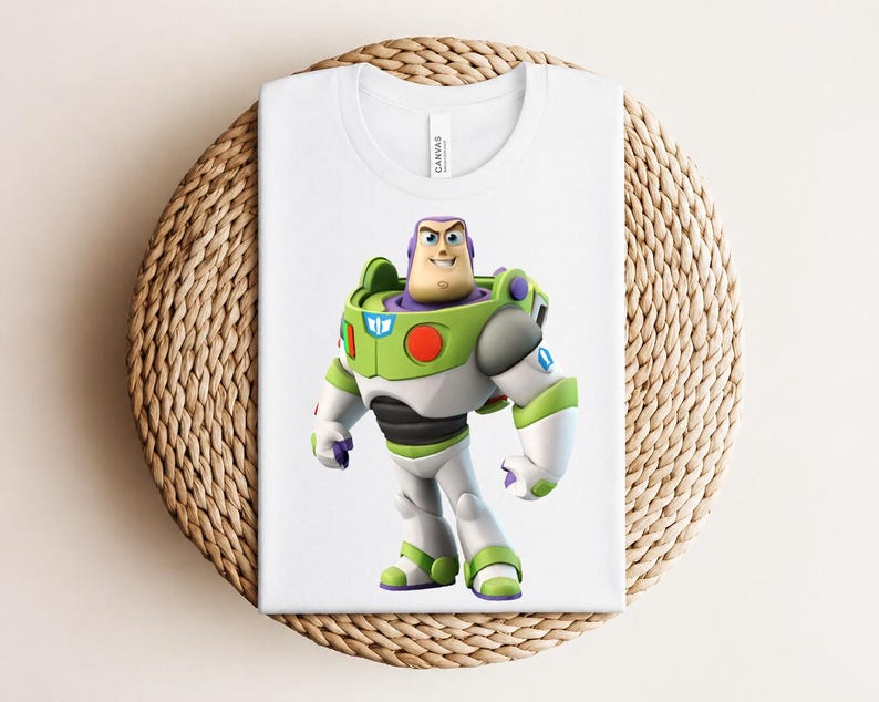May include: White t-shirt featuring a colorful graphic of Buzz Lightyear from Toy Story. The character is depicted in his signature green, white, and purple spacesuit. The shirt is folded on a woven, circular surface.