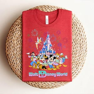 Może przedstawiać: Czerwona koszulka z kolorową grafiką postaci Disneya, w tym Myszki Miki i Minnie, Kaczora Donalda i Goofiego, na tle zamku i fajerwerk&oacute;w. Tekst "Walt Disney World" jest wyświetlany poniżej.