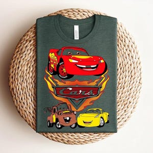 May include: Dark green t-shirt featuring a graphic of characters from the animated movie "Cars." The design includes a red race car, a brown truck, and a yellow car, with the word "Cars" in a stylized font.