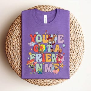 May include: A lilac t-shirt with the words "You've Got a Friend in Me" in colourful, retro lettering. The shirt features cartoon characters from a popular animated film. The t-shirt is displayed on a woven, circular surface.