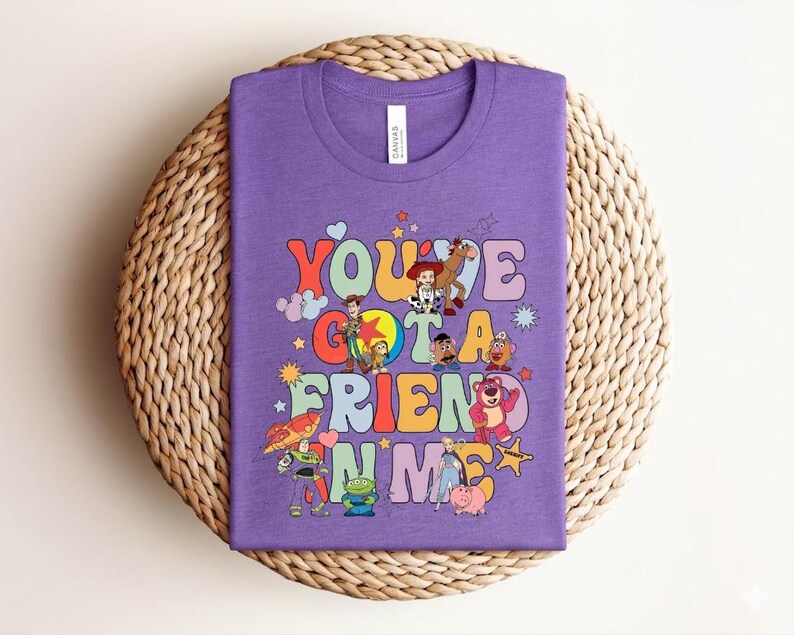 May include: A lilac t-shirt with the words "You've Got a Friend in Me" in colourful, retro lettering. The shirt features cartoon characters from a popular animated film. The t-shirt is displayed on a woven, circular surface.
