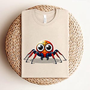 Pu&ograve; includere: T-shirt beige con un design a ragno a cartoni animati colorato. Il ragno ha un corpo rotondo con sfumature gialle, arancioni e rosse, grandi occhi e zampe rosse e blu. La maglietta &egrave; piegata su una superficie tessuta.