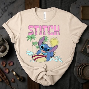 May include: Beige t-shirt featuring a graphic of Stitch surfing on a surfboard. The word "STITCH" is printed above the character in pink block letters. Palm trees and a yellow sun are also in the design.